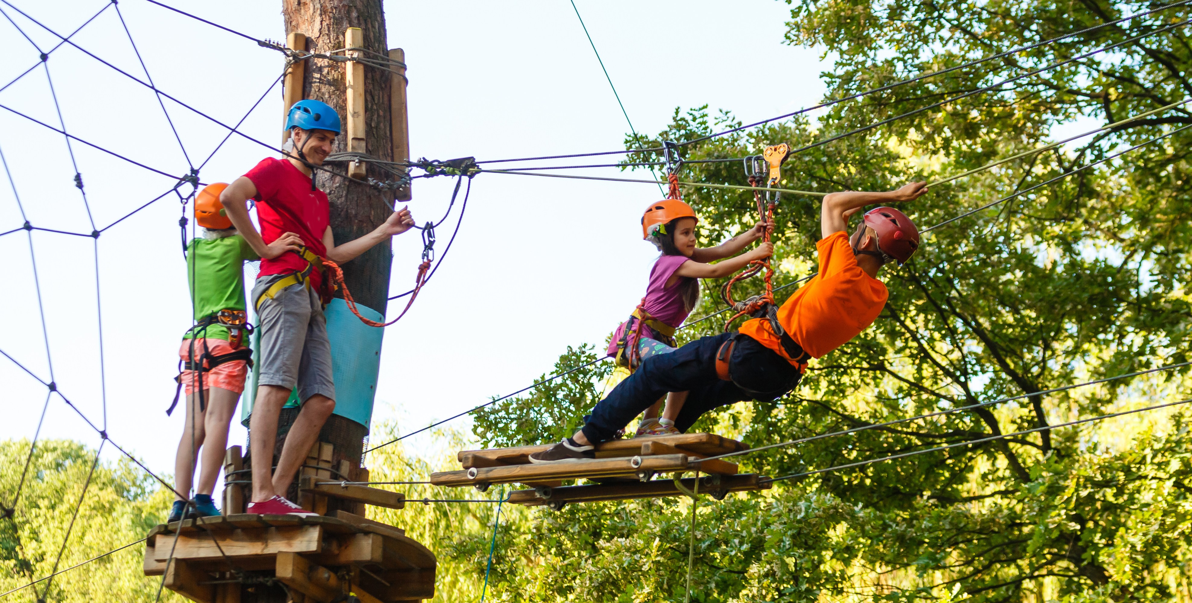 People with helmets zip line on a forest ropes course on a sunny day.