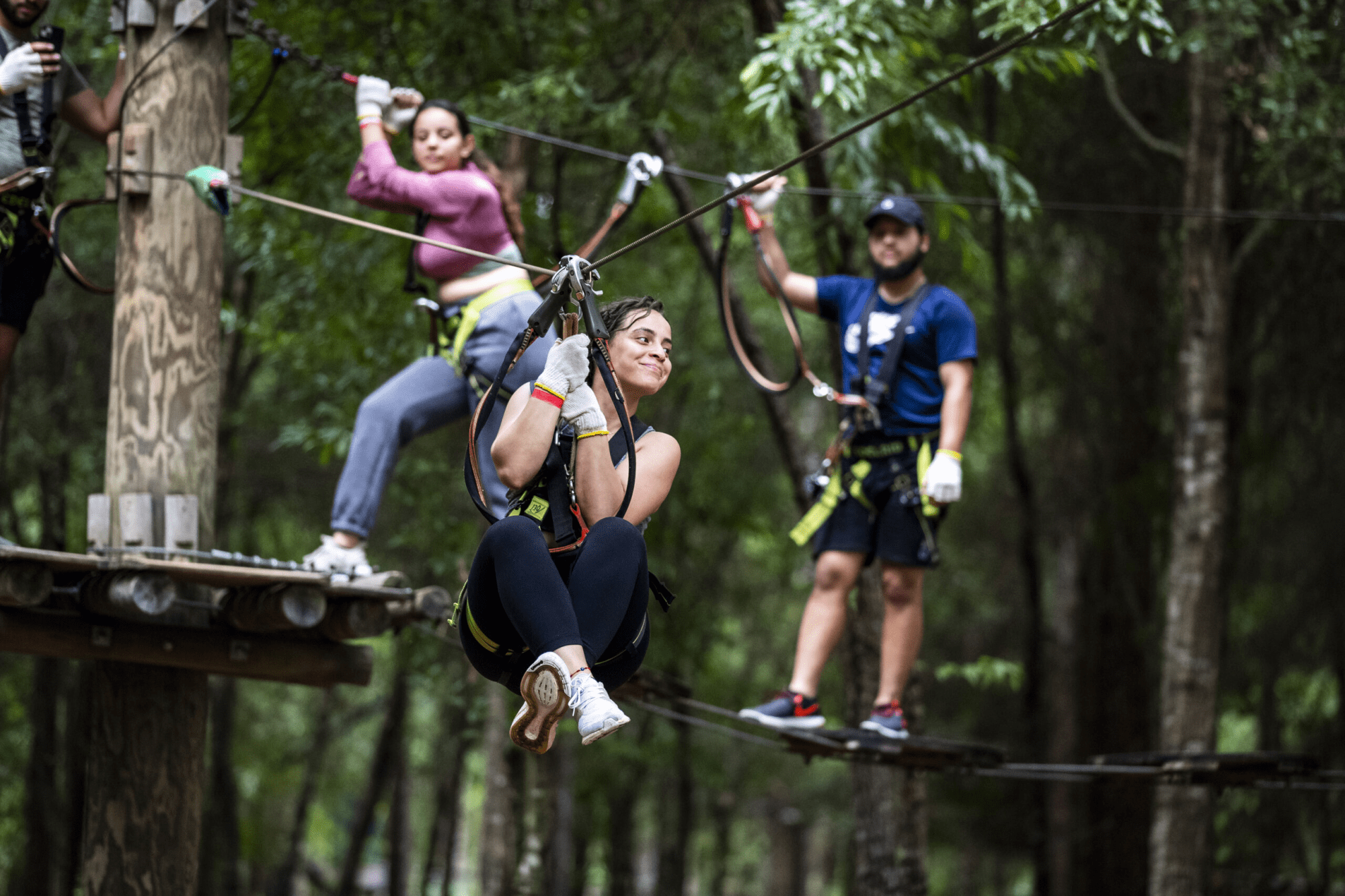 Three people zip-lining in a forest, smiling and wearing safety gear.