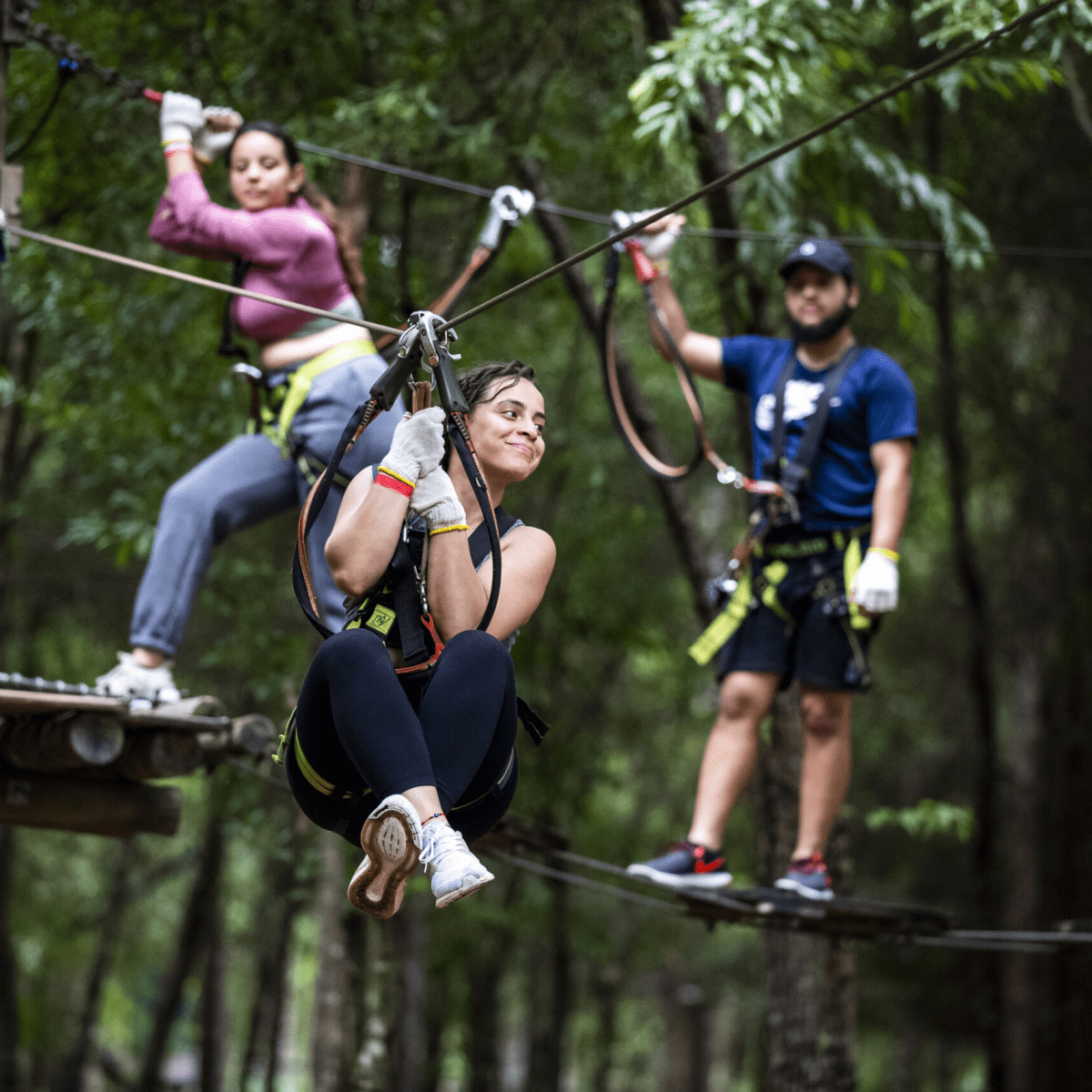 Three people zip-lining in a forest, smiling and wearing safety gear.