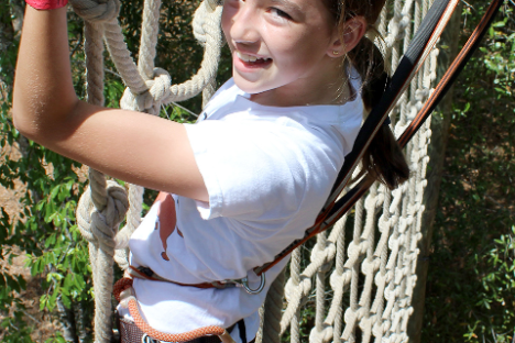 Girl in a white shirt and harness climbing a rope net outdoors.