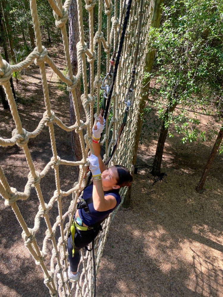 Person climbing a vertical rope net in a forest, wearing harness and gloves.