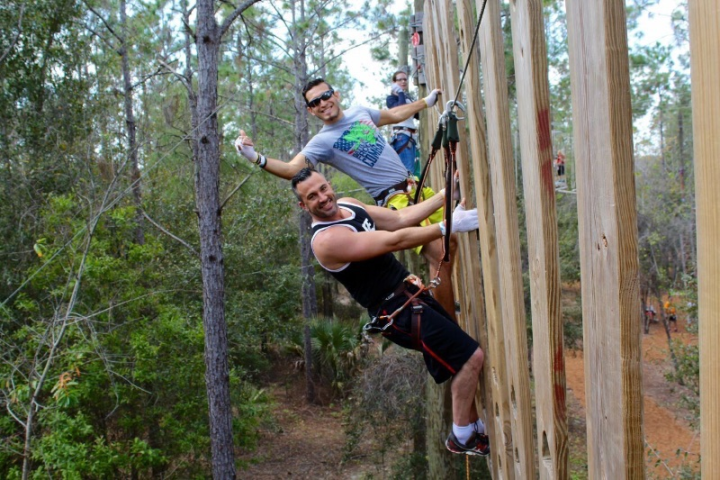 Two people climbing a wooden wall in a forested area, smiling at the camera.