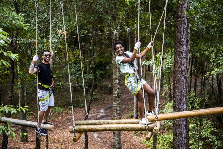 Two people on a rope bridge in a forest, smiling and wearing safety harnesses.
