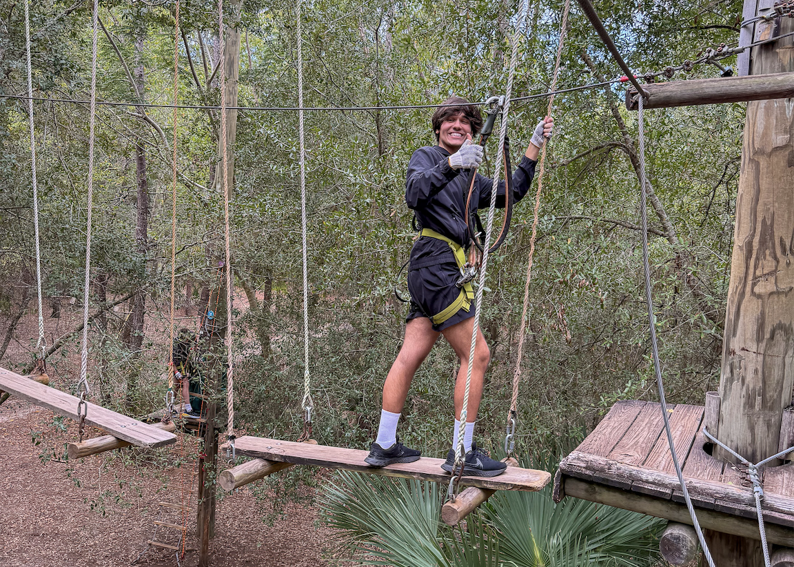 Person on outdoor ropes course bridge, wearing harness, surrounded by trees.